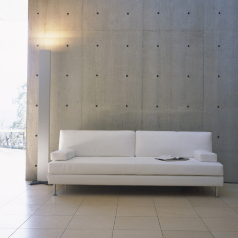 株式会社アイデック PLATEAUのソファ。打ち放しコンクリートの壁面に沿って配置された、白いクッションと金属製の円柱状の脚で構成された3人掛けソファ。左側には垂直な板状のフロアランプが置かれ、コンクリート面を上方から照らしている構図。